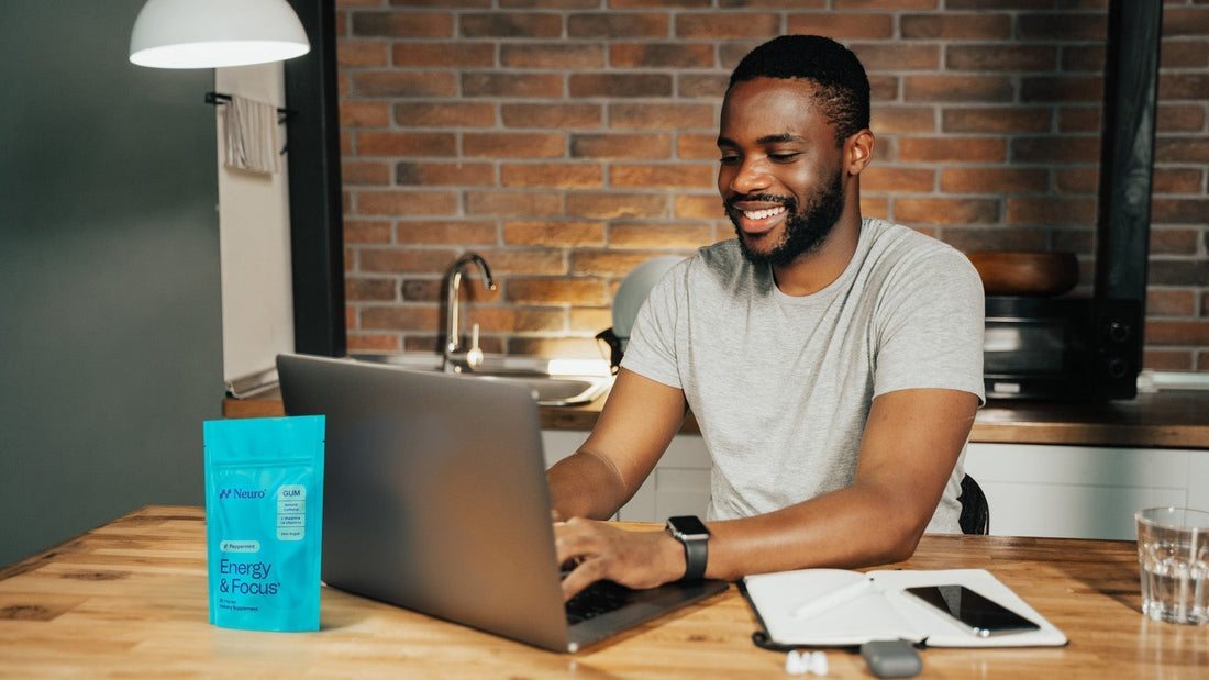 A man working from home, typing on his computer with a packet of Neuro Energy and Focus gum on his desk